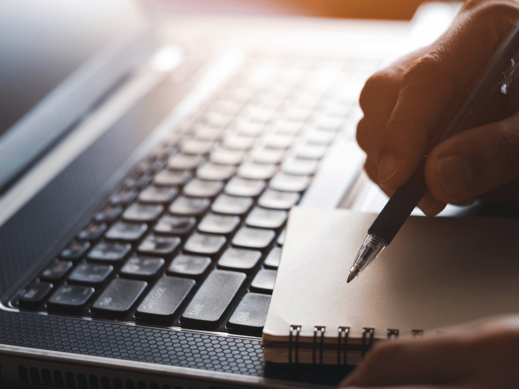 Egy üres füzet, amibe éppen ír egy ember tollal, háttérben egy laptop billentyűzete - webinár felkészülés